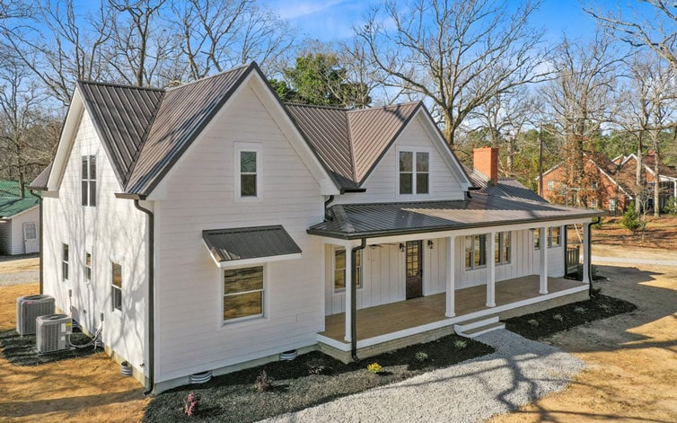 Beautiful white farmhouse with a large wraparound porch and black roof, set in a wooded area.l